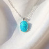 A silver chain necklace with an oval turquoise-blue opal pendant, resting on a white fabric surface.