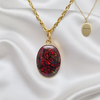 A gold chain necklace with an oval pendant containing red and green iridescent glitter, displayed on white satin fabric next to a gold oval tag engraved with 'Semperey'.