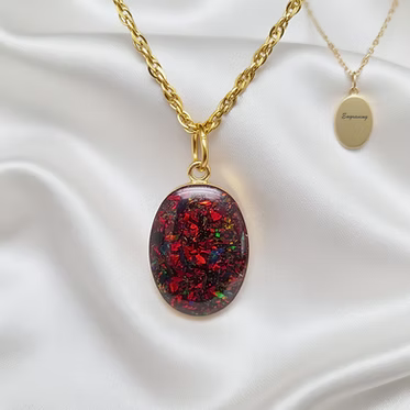 A gold chain necklace with an oval pendant containing red and green iridescent glitter, displayed on white satin fabric next to a gold oval tag engraved with 'Semperey'.