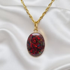 An oval pendant necklace with a gold chain, featuring a dark red and black opal-like resin pendant filled with iridescent glitter, displayed on a white satin fabric background.