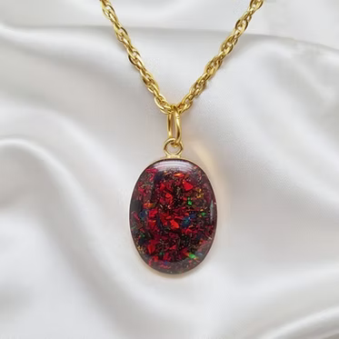 An oval pendant necklace with a gold chain, featuring a dark red and black opal-like resin pendant filled with iridescent glitter, displayed on a white satin fabric background.