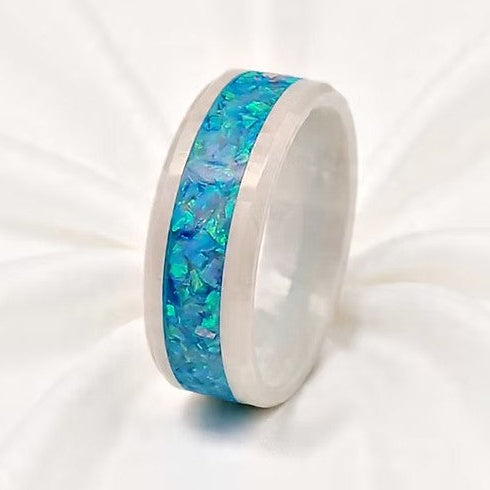 A polished white ceramic band ring with a vibrant blue and green iridescent inlay, displayed on a soft white fabric background.
