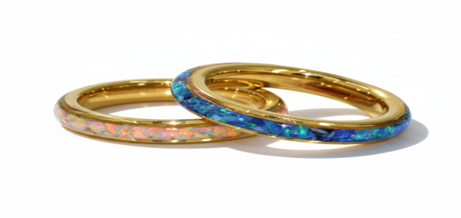 Two gold rings with inlaid opal, one showing blue and green flashes and the other pink and orange flashes, resting on a white satin fabric.