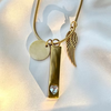 Gold-toned necklace with a snake chain, a flat round disc charm, a wing charm, and a vertical bar pendant featuring a clear heart-shaped crystal at the bottom, displayed on a white fabric background.