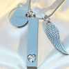 Silver-toned cremation urn necklace with a vertical bar pendant featuring a heart-shaped crystal, a wing charm, and a round disc pendant on a silver chain, displayed on a white satin background.