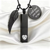 Black cremation urn necklace featuring a vertical rectangular pendant with a heart-shaped clear stone, a black angel wing charm, and a round engraved tag that reads 'I still need you close to me.' displayed on white satin fabric.