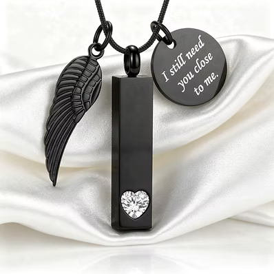 Black cremation urn necklace featuring a vertical rectangular pendant with a heart-shaped clear stone, a black angel wing charm, and a round engraved tag that reads 'I still need you close to me.' displayed on white satin fabric.