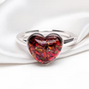 A silver band ring featuring a heart-shaped clear resin cabochon filled with red and orange dried flower petals, resting on a white satin fabric.