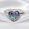 A silver heart-shaped ring featuring an iridescent abalone shell inlay with shimmering purple, green, and blue tones, displayed on a soft white satin fabric.
