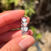 A close-up of a hand holding a silver-plated brass alloy ring with a row of iridescent heart-shaped opal stones against a blurred outdoor background.
