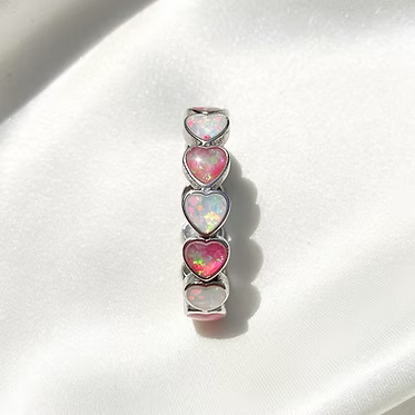 A silver-toned ring featuring five stacked heart-shaped inlays with pink and iridescent opal-like finishes, displayed on a white satin fabric background.