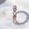A silver-plated brass alloy ring featuring five stacked heart-shaped opal stones in pink, white, and iridescent colors, displayed on a soft white fabric background.