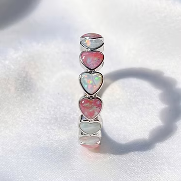 A silver-plated brass alloy ring featuring five stacked heart-shaped opal stones in pink, white, and iridescent colors, displayed on a soft white fabric background.
