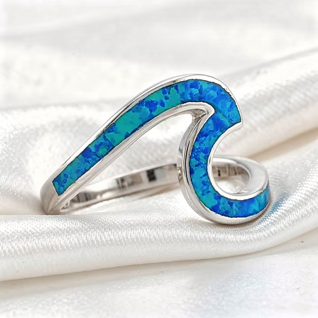 A silver wave-shaped ring featuring vibrant blue and turquoise inlay, displayed on a white satin fabric background.