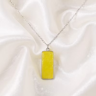 A silver chain necklace with a rectangular yellow pendant, displayed on draped white satin fabric.