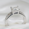 A silver solitaire ring with a rectangular clear gemstone, resting on a white satin fabric background.