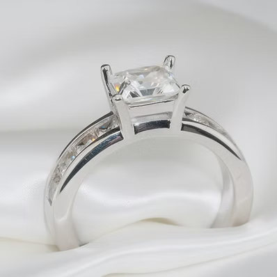 A silver solitaire ring with a rectangular clear gemstone, resting on a white satin fabric background.