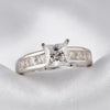 A silver engagement ring featuring a princess-cut clear center stone and a channel-set row of small clear stones on the band, displayed on a white textured fabric surface.