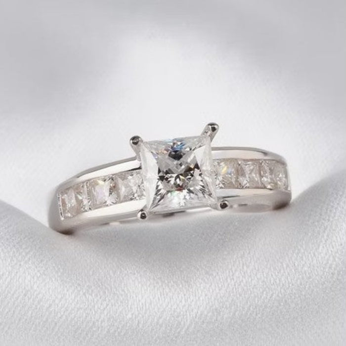 A silver engagement ring featuring a princess-cut clear center stone and a channel-set row of small clear stones on the band, displayed on a white textured fabric surface.