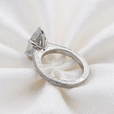 A silver ring with an engraved band and a large clear gemstone, displayed on a textured white fabric background.