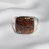 A silver-toned signet ring featuring a square cabochon gemstone with a highly iridescent, glittery opal-like pattern of orange, green, red, and black flecks, displayed on a soft white fabric background.