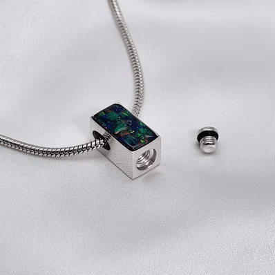 A silver snake chain necklace with a rectangular silver charm featuring a colorful iridescent inlay, shown next to its small silver end cap on a white textured fabric background.