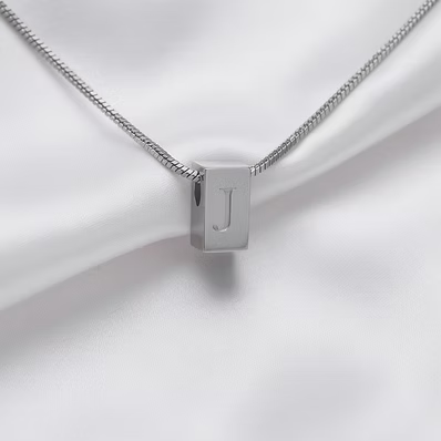 A silver box pendant necklace with a polished letter 'J' on a silver snake chain, displayed on a draped white satin fabric.