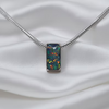 A silver snake chain necklace with a rectangular pendant featuring a dark, iridescent opal-like gemstone with vibrant orange, yellow, and red flashes, displayed on a draped white satin fabric.