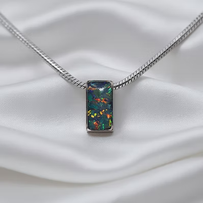 A silver snake chain necklace with a rectangular pendant featuring a dark, iridescent opal-like gemstone with vibrant orange, yellow, and red flashes, displayed on a draped white satin fabric.