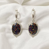A pair of silver leverback earrings with oval iridescent opal-like stones, displayed on a soft white fabric background.