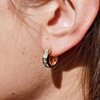 A close-up of a person's ear wearing a small gold huggie hoop earring with a black enamel inlay and iridescent square accents.