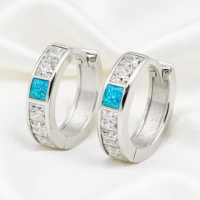 A pair of silver huggie hoop earrings with clear rhinestone accents and a central square blue stone, displayed on a soft white satin background.