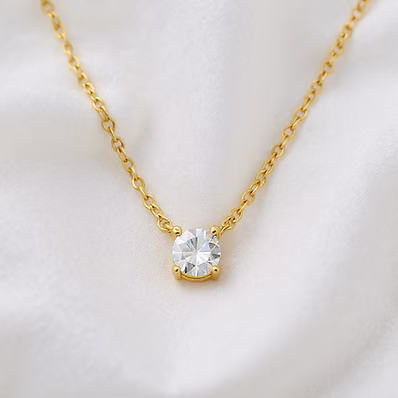 A delicate gold chain necklace with a single round clear gemstone pendant, displayed on a soft white fabric background.