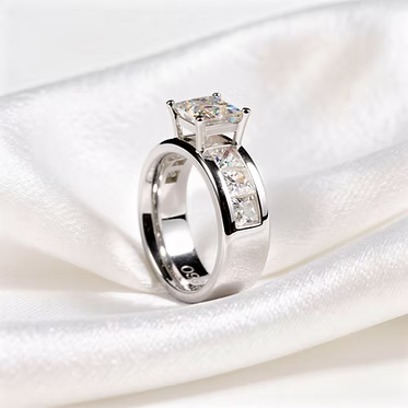 A silver-toned ring with a square-cut clear center stone and a row of small clear stones along the band, displayed on white satin fabric.