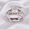 A silver-plated princess-cut diamond engagement ring with a matching diamond band, resting on a draped white satin fabric.