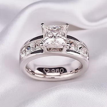 A silver-plated princess-cut diamond engagement ring with a matching diamond band, resting on a draped white satin fabric.