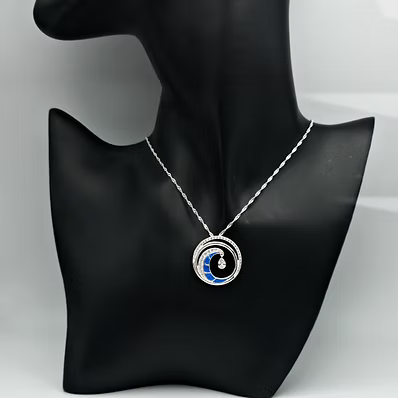 A silver-toned necklace with a circular pendant featuring concentric rings and a blue crescent moon design, displayed on a black jewelry bust against a light gray background.