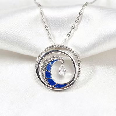A silver-toned crescent moon pendant necklace with a deep blue enamel center, surrounded by small clear stones, resting on a white satin fabric background.