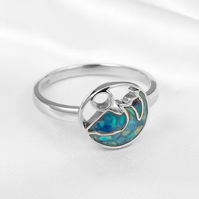 An argentium silver ring featuring a circular bezel-set cabochon with a vibrant blue and green opal inlay, displayed on a soft white fabric background.