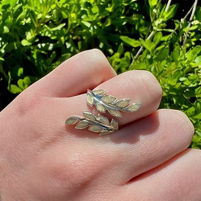 A silver leaf-shaped ring worn on a person's finger, with a small blue stone or inlay, set against a background of green outdoor foliage.