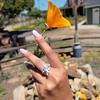 A close-up lifestyle shot of a hand wearing a silver ring with a leaf-like design, holding a bright orange poppy flower outdoors with a blurred rustic background.