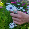 A hand wearing a silver ring decorated with green and blue enamel leaves, holding a white and purple daisy flower against a blurred background of green foliage and colorful wildflowers.