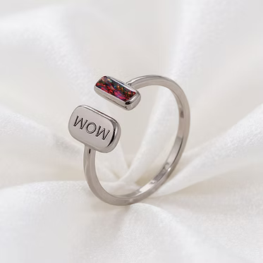 A silver-toned open ring band featuring a polished rectangular silver charm engraved with 'WOW' and a smaller top bar with a colorful, iridescent inlay, displayed on a soft white fabric background.