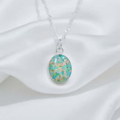 An oval pendant necklace with a vibrant turquoise, green, and gold opal-like stone set in a silver bezel, hanging from a silver chain against a draped white satin background.