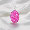 An oval pendant necklace with a vibrant pink glittery resin center and a silver-toned chain, displayed on a draped white satin fabric background.