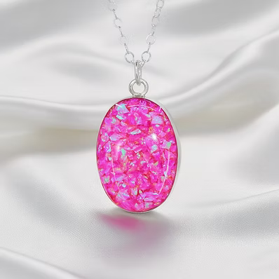 An oval pendant necklace with a vibrant pink glittery resin center and a silver-toned chain, displayed on a draped white satin fabric background.
