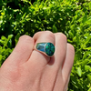 A sterling silver signet ring with a textured green stone, worn on a hand with a background of green foliage.