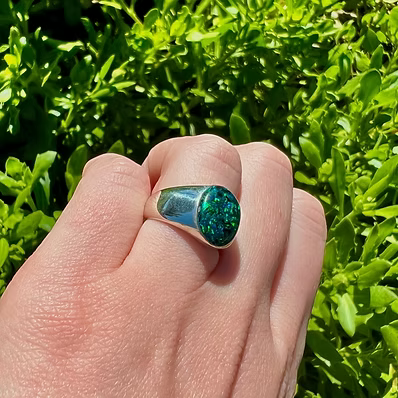 A sterling silver signet ring with a textured green stone, worn on a hand with a background of green foliage.