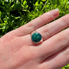 A silver ring with a round, glittery teal-green cabochon gemstone worn on a person's finger, set against a background of bright green foliage.