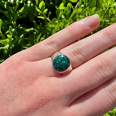 A silver ring with a round, glittery teal-green cabochon gemstone worn on a person's finger, set against a background of bright green foliage.